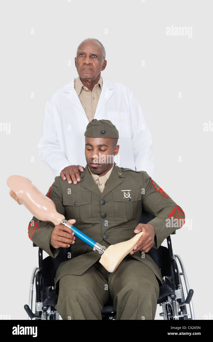 Leitender Arzt mit US-Offizier hält künstlichen Gliedmaßen, da er im Rollstuhl über den grauen Hintergrund sitzt Stockfoto