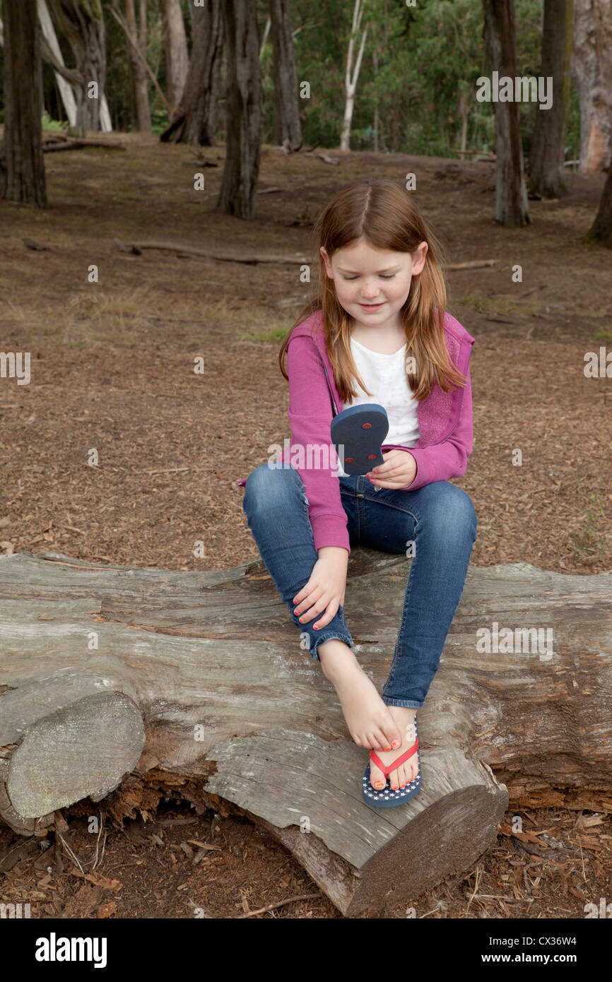 Kleines Mädchen sitzt auf einem umgestürzten Baum mit einem Flip flop in der Hand. Stockfoto