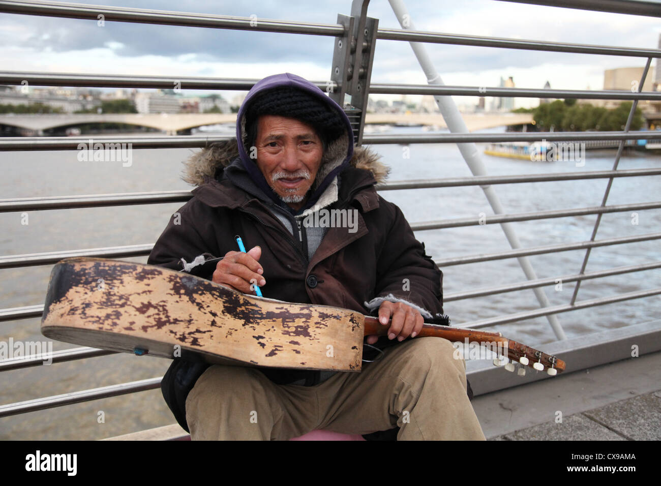 Ein Obdachloser zupft eine Zeichenfolge auf seiner Gitarre, während er Passanten für den Wandel auf Hungerford Bridge London fragt. Stockfoto