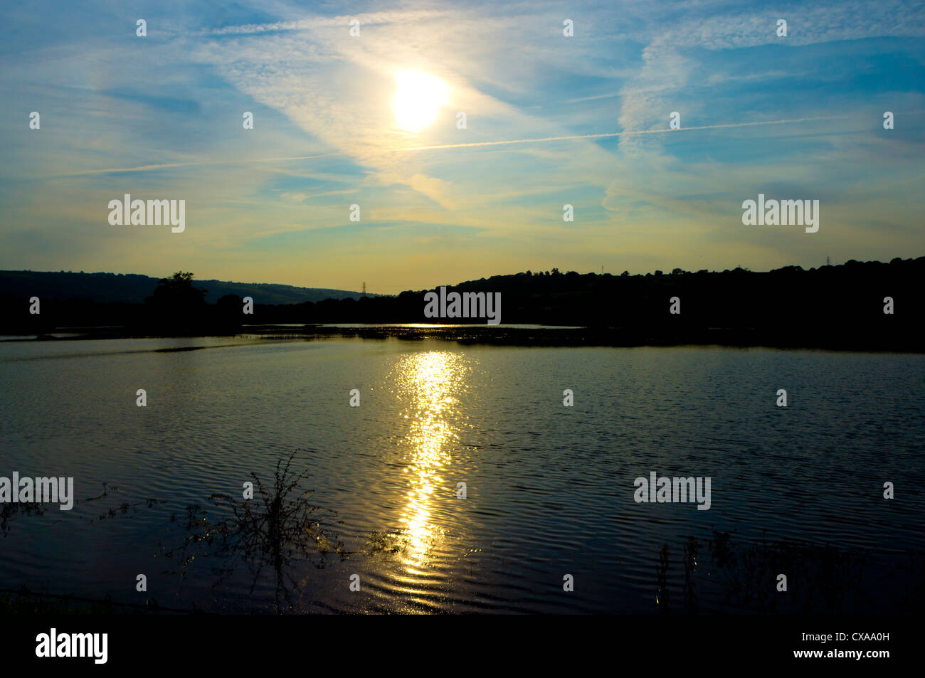 Die Sonne über einem See unter einem Dampf-Trail gestreift Himmel setzt, und erstellt eine Welle von Sonnenschein über das Wasser. Stockfoto