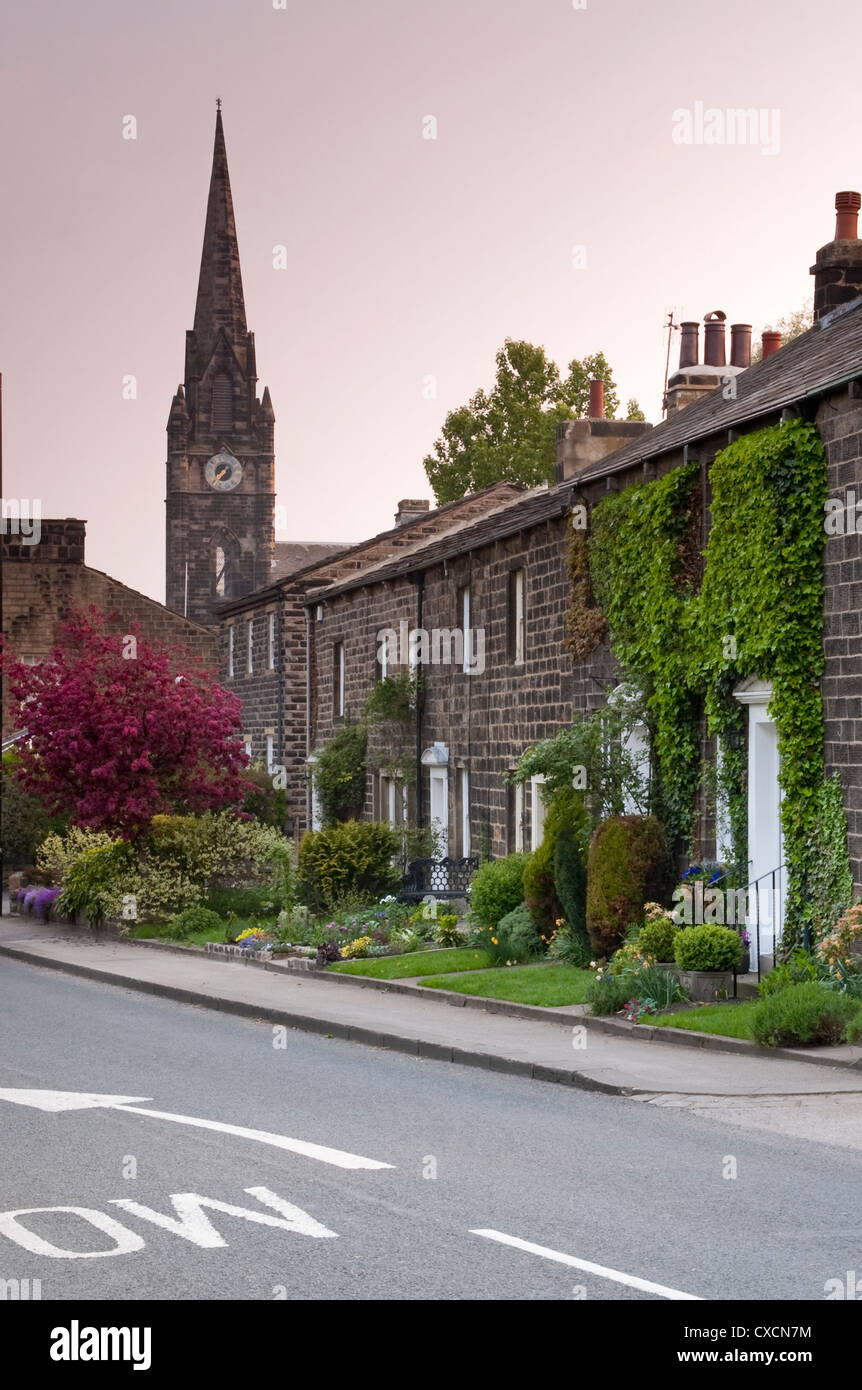 Terrasse Zeile aus Stein gebauten Cottages am Straßenrand mit hoch aufragenden Turm & Uhr der St. Mary's Kirche jenseits - Burley-in-Wharfedale, Yorkshire, England, UK. Stockfoto