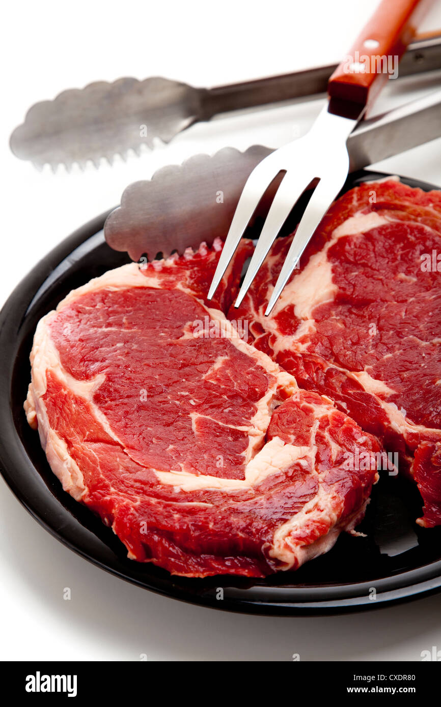 Eine Gruppe von rohes Rindfleisch Ribeye Steaks auf einem weißen Hintergrund mit Bbq oder Kochutensilien, einschließlich eine Gabel und eine Zange Stockfoto