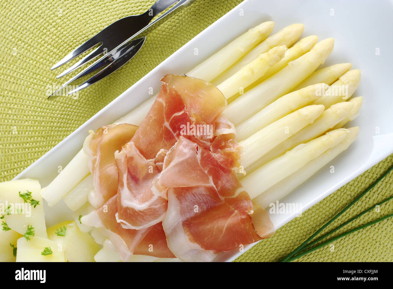 Frisch gekochter weißer Spargel mit Schinken und Kartoffeln auf Langschild mit Schnittlauch und Besteck an den Seiten serviert Stockfoto