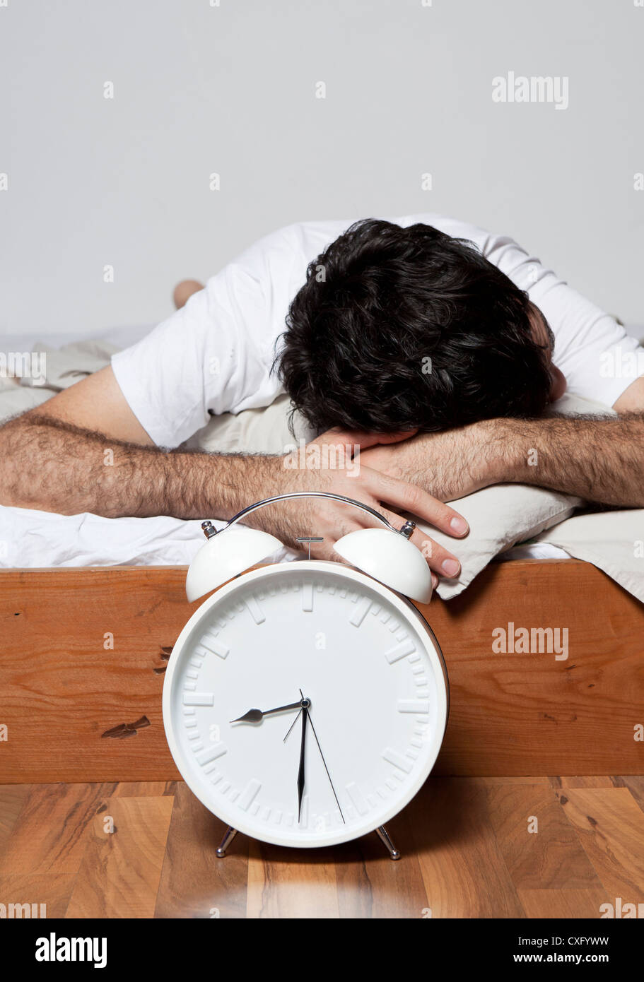 Ein junger Mann schläft auf einem Bett in der Nähe von einem großen weißen Wecker. Stockfoto