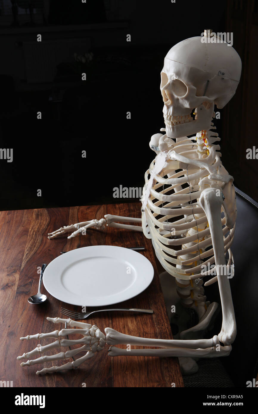 Skelett sitzen am gedeckten Tisch mit einer Platte, ein Messer, einen Löffel und einer Gabel Stockfoto