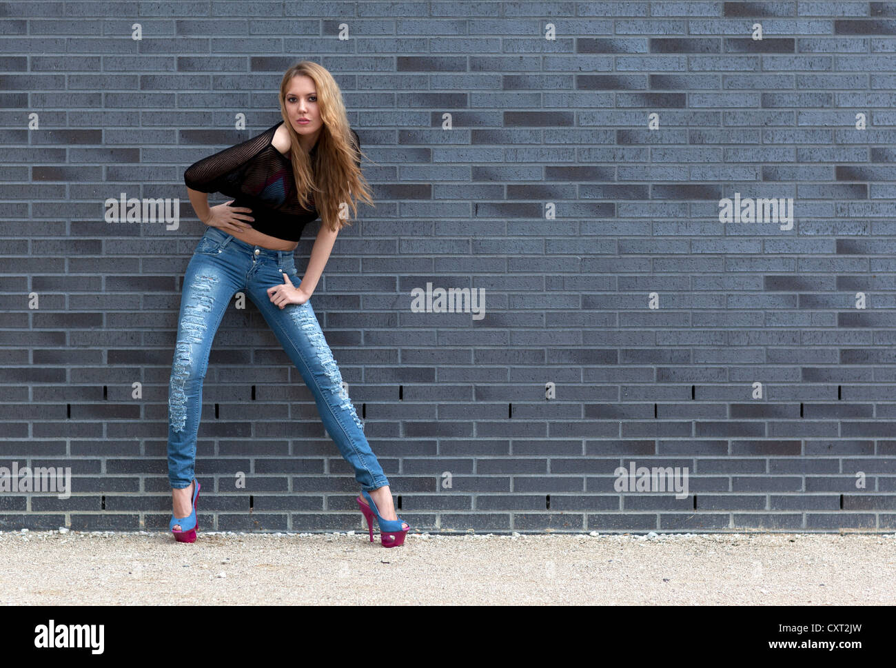 Junge Frau in zerrissenen Jeans, schwarzes Top und high Heels posiert vor einer Wand Stockfoto