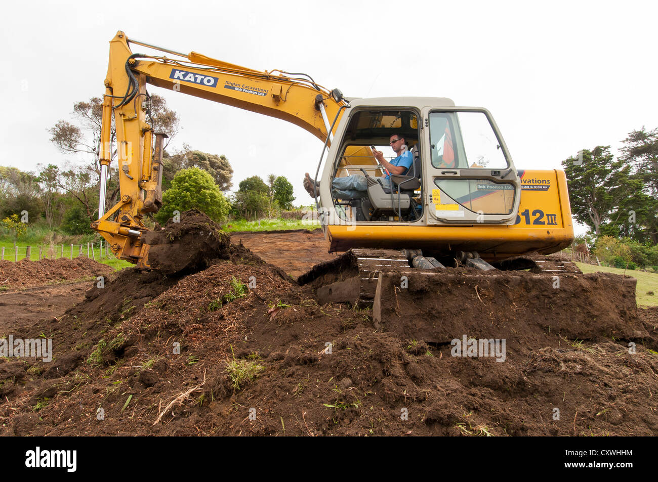 Mann sitzt im Inneren Digger am Telefon, beim Ausheben Boden für neue Heimat am steil abfallenden Abschnitt, umgeben von einer ländlichen Landschaft. Stockfoto