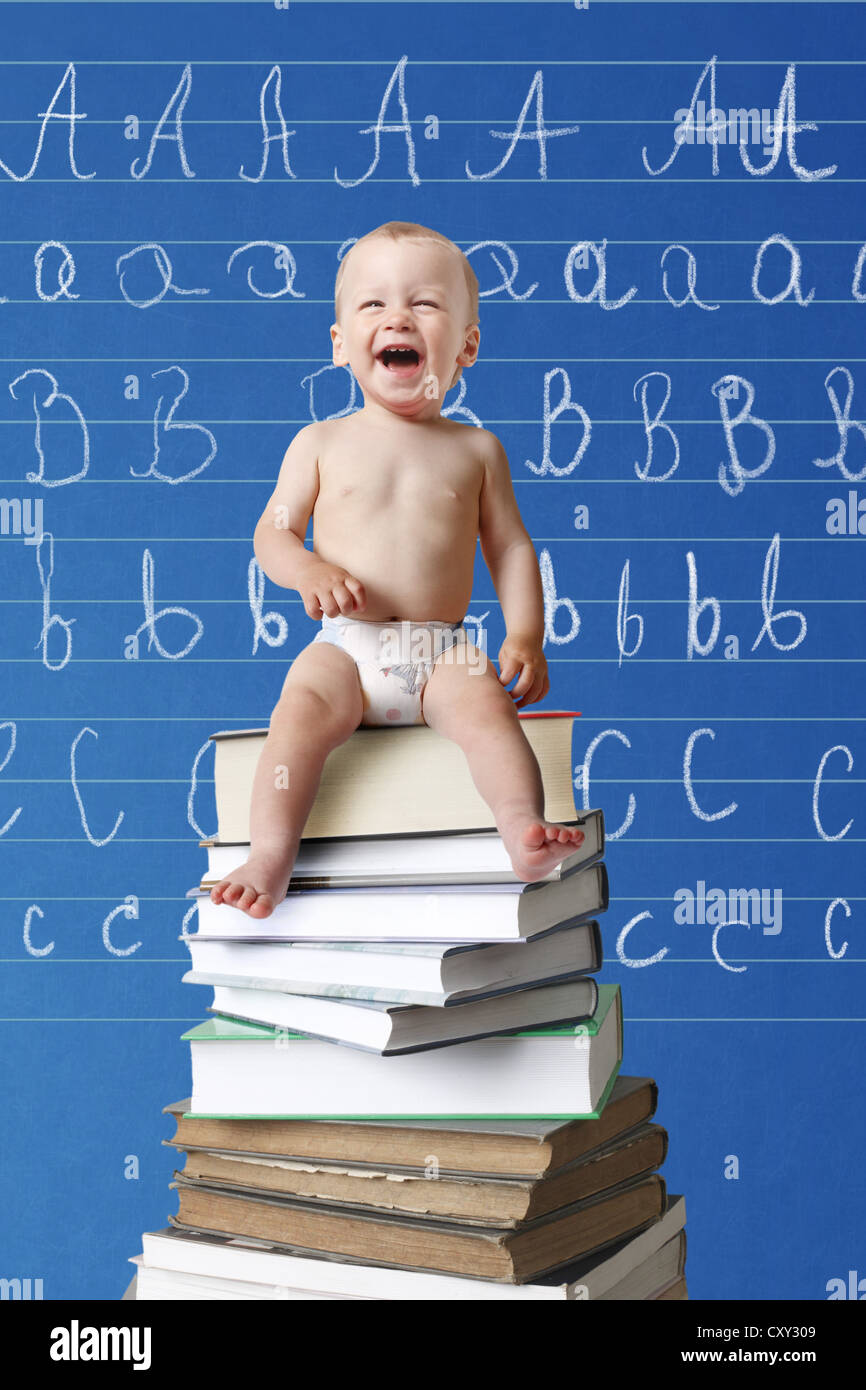 Lachendes Baby sitzt auf einem Stapel von Büchern vor einer Tafel mit Buchstaben darauf geschrieben Stockfoto