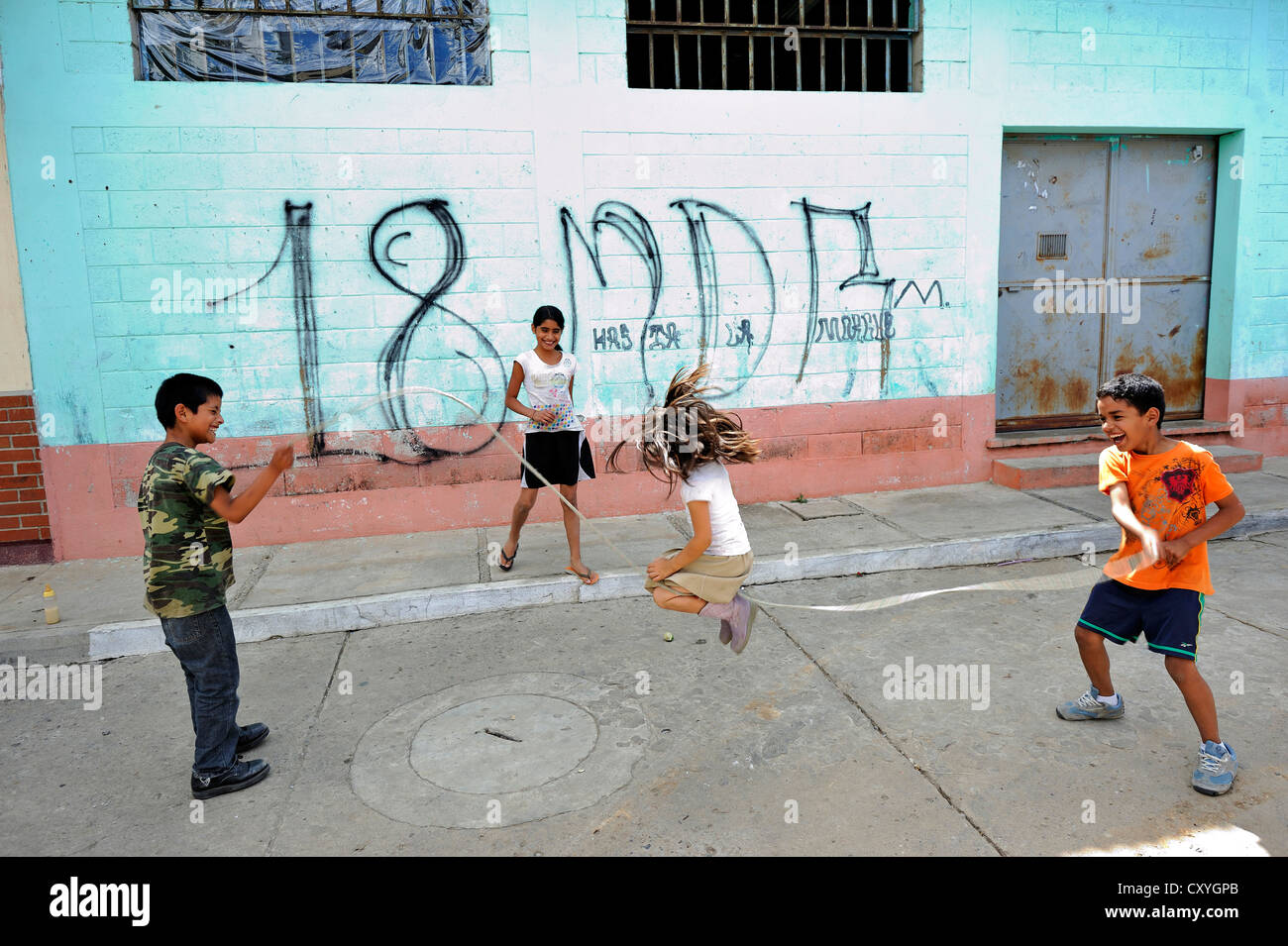 Kinder spielen vor einer Wand mit Graffiti der Mara 18-Bande von Jugendlichen, Lomas de Santa Faz slum Stockfoto