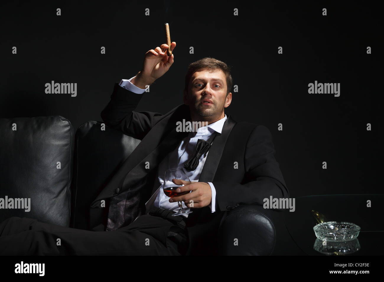Macho Mann in einem stilvollen Tuxedo sitzen in der Dunkelheit in einem Nachtclub schnaufend auf einer Zigarre und trinken Weinbrand oder cognac Stockfoto