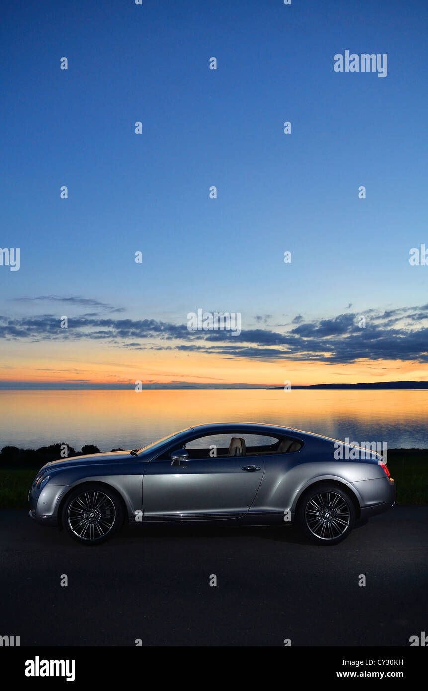 Bentley Continental GT neben Strand und Meer mit Sonnenuntergang und Inseln im Hintergrund und Wolken im Himmel Stockfoto