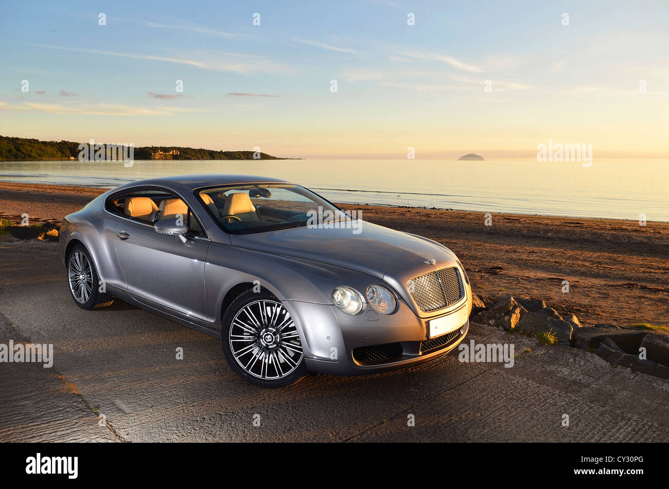 Bentley Continental GT neben Strand und Meer mit Sonnenuntergang und Inseln im Hintergrund und Wolken im Himmel Stockfoto