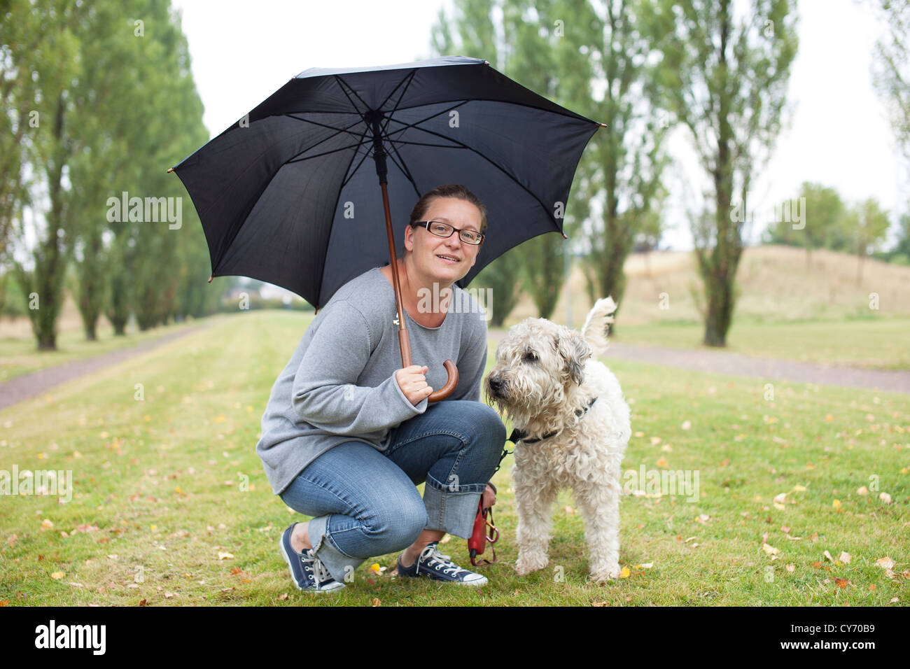 Frau und ihr Hund unter einem Regenschirm. Sie sucht mit einem freundlichen Lächeln in die Kamera. Kleidung ist leger. Stockfoto