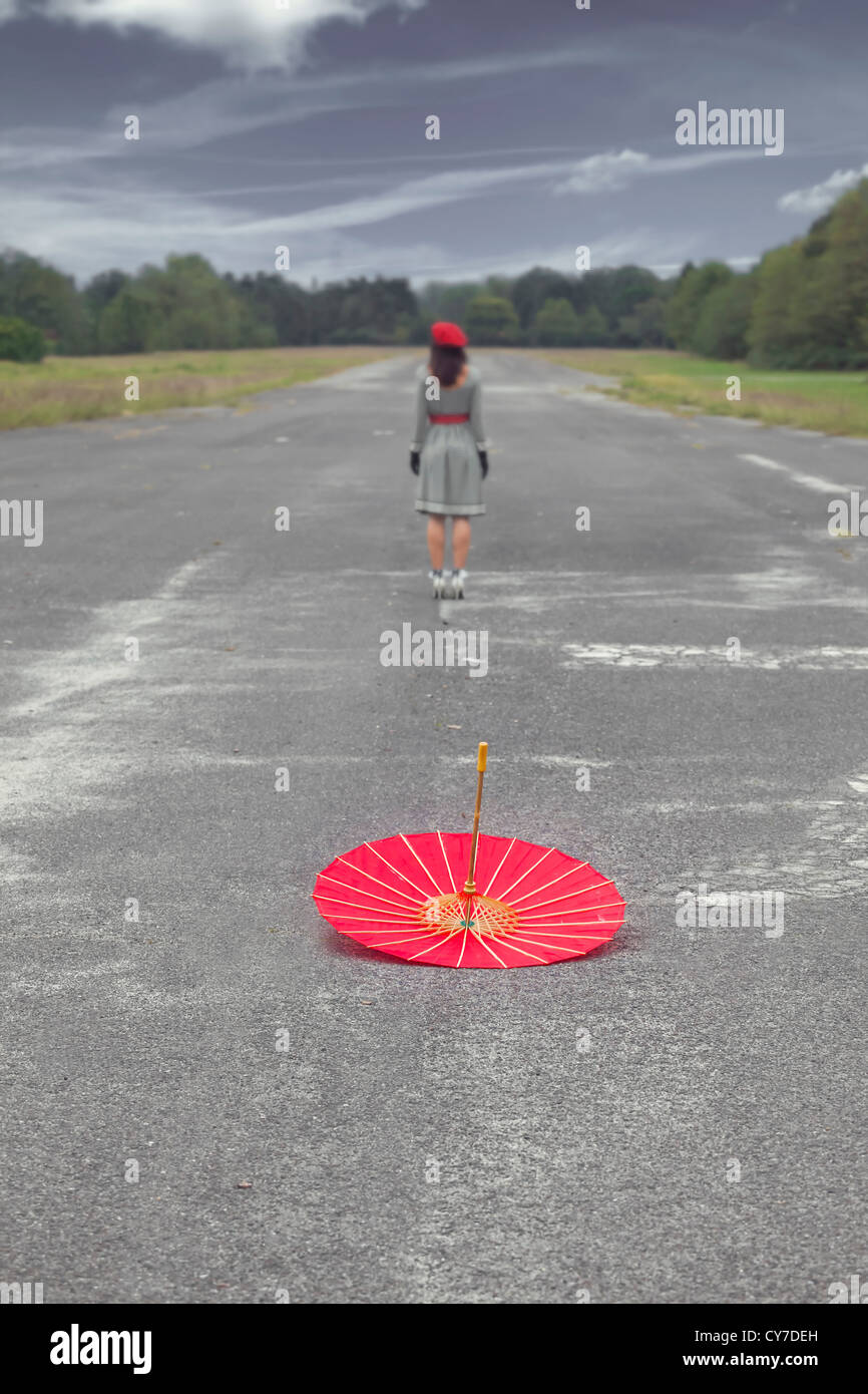 ein roter Regenschirm auf einer Straße liegt, eine Frau steht in einer Entfernung Stockfoto