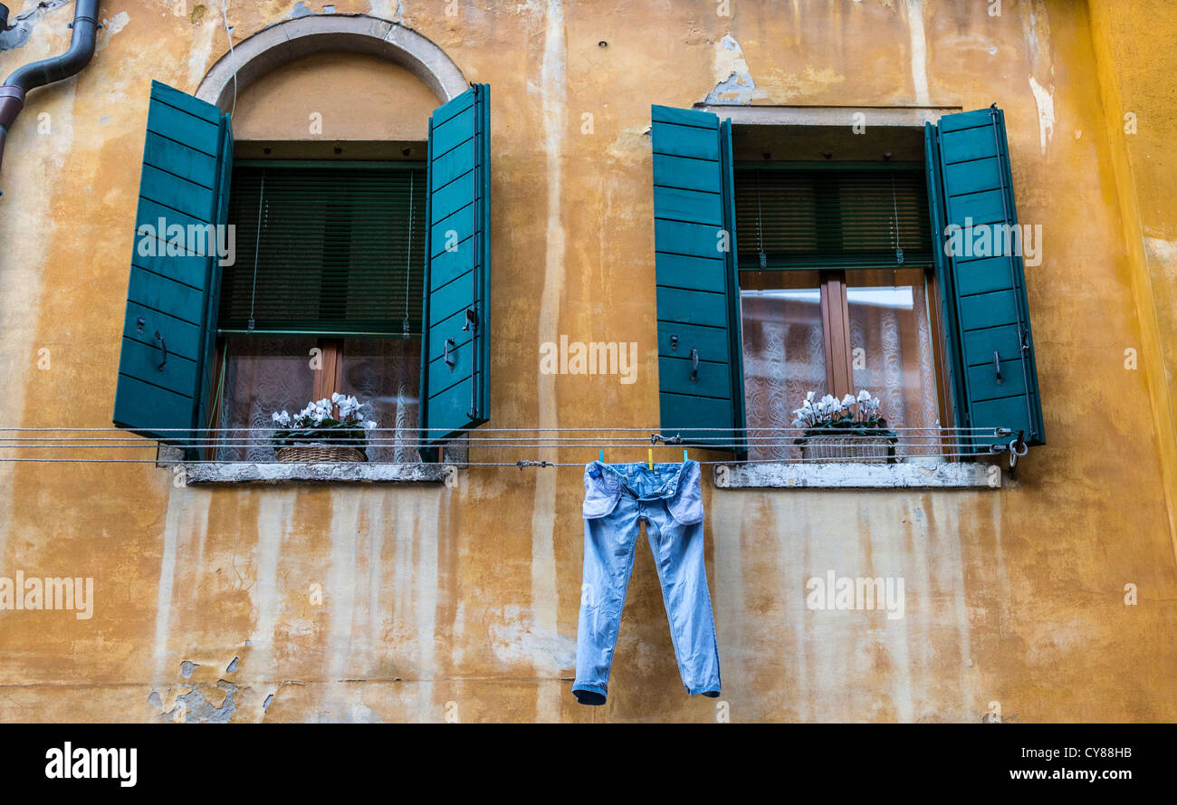 Verblasst und gefärbt gelb-ocker-Wand, Fenster mit blauen Fensterläden und ein paar blaue Denimjeans hängen zum Trocknen in Venedig Stockfoto