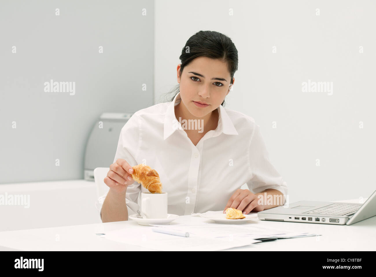 Junge Frau eintauchen Croissant in Kaffee am Schreibtisch im Büro Stockfoto
