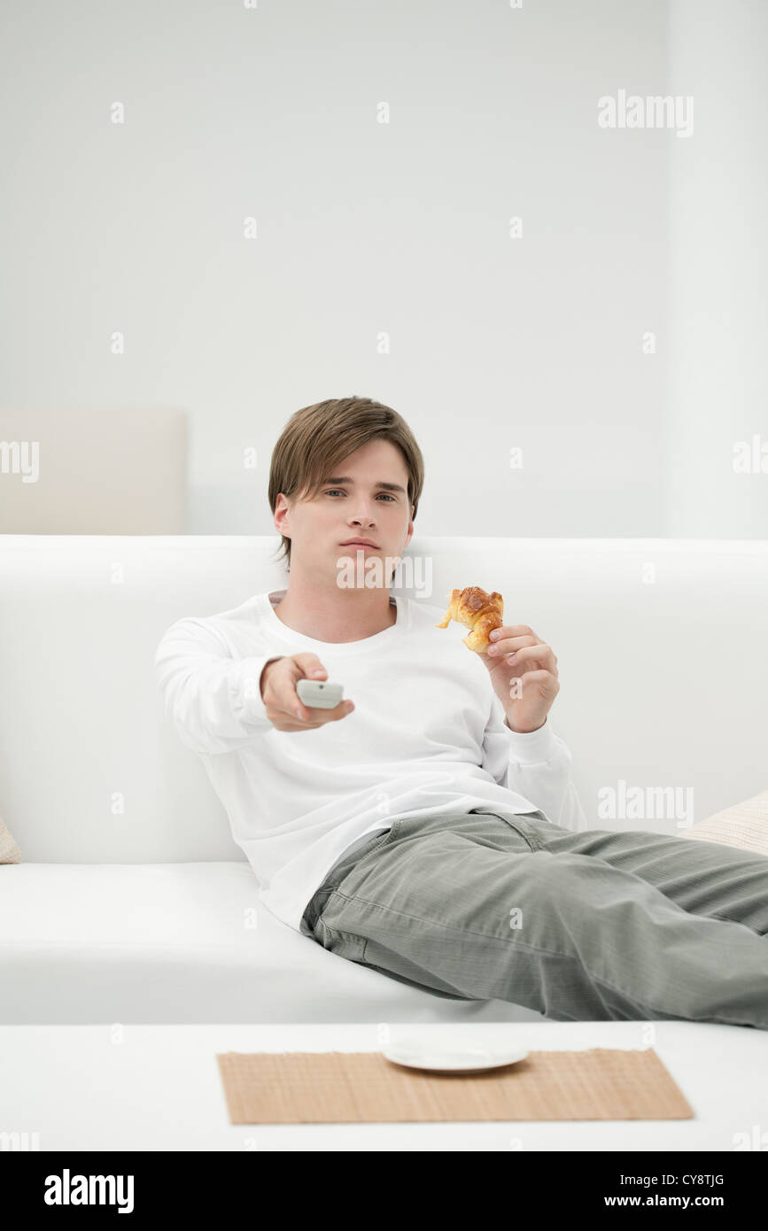 Junger Mann isst Croissant beim Fernsehen Stockfoto