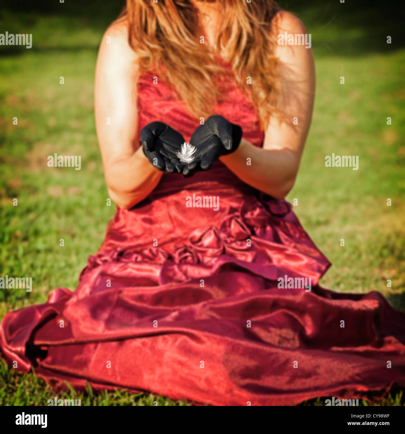 eine Frau in einem roten Kleid sitzt auf einer Wiese, eine Feder in ihren Händen hält Stockfoto