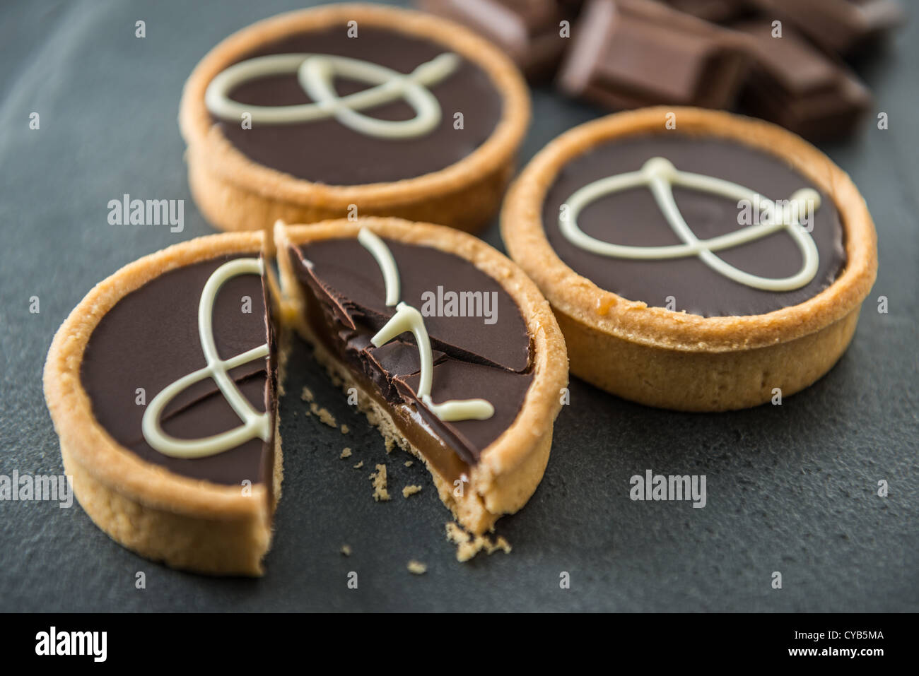 Drei Schokolade Törtchen im Vordergrund mit Schokoladenstücken im Hintergrund auf einem Schieferoberfläche. Eine Torte wurde gekürzt. Stockfoto