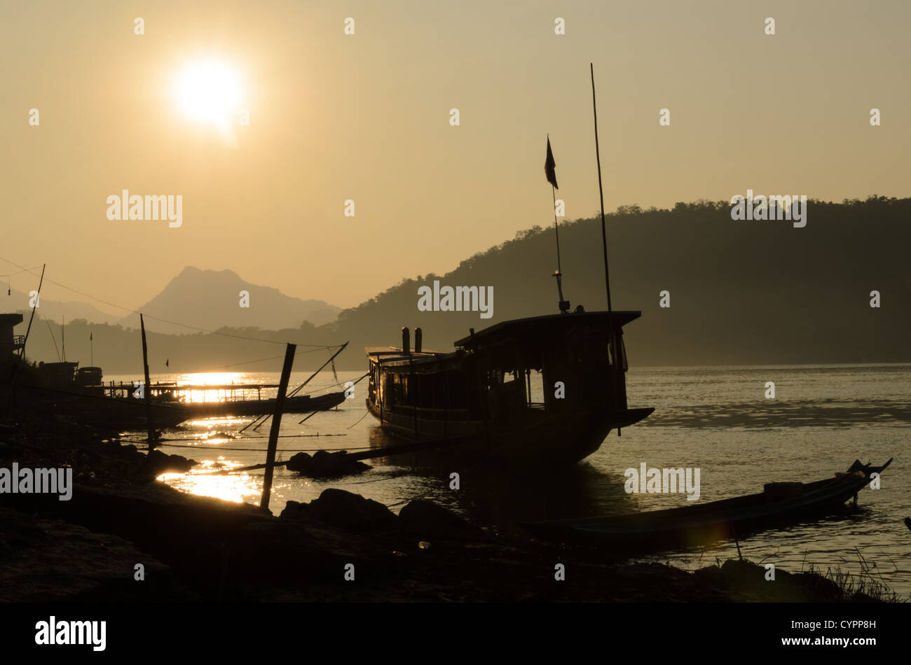 Sonnenuntergang Luang Prabang Laos // LUANG PRABANG, Laos — Ein hölzernes, überdachtes Kabinenboot ist vor der untergehenden Sonne auf dem Mekong in der Nähe von Luang Prabang im Norden Laos gehalten. Der Fluss Mekong dient als wichtige Transportroute durch diese UNESCO-Weltkulturerbe-Stadt und verbindet die Gemeinden entlang der Ufer. Luang Prabang liegt am Zusammenfluss der Flüsse Mekong und Nam Khan in Nord-Zentral-Laos. Traditionelle Holzboote wie diese bleiben ein wichtiges Transport- und Handelsmittel entlang des Flusses. Die alte königliche Hauptstadt ist bekannt für ihre gut erhaltene Architektur Stockfoto