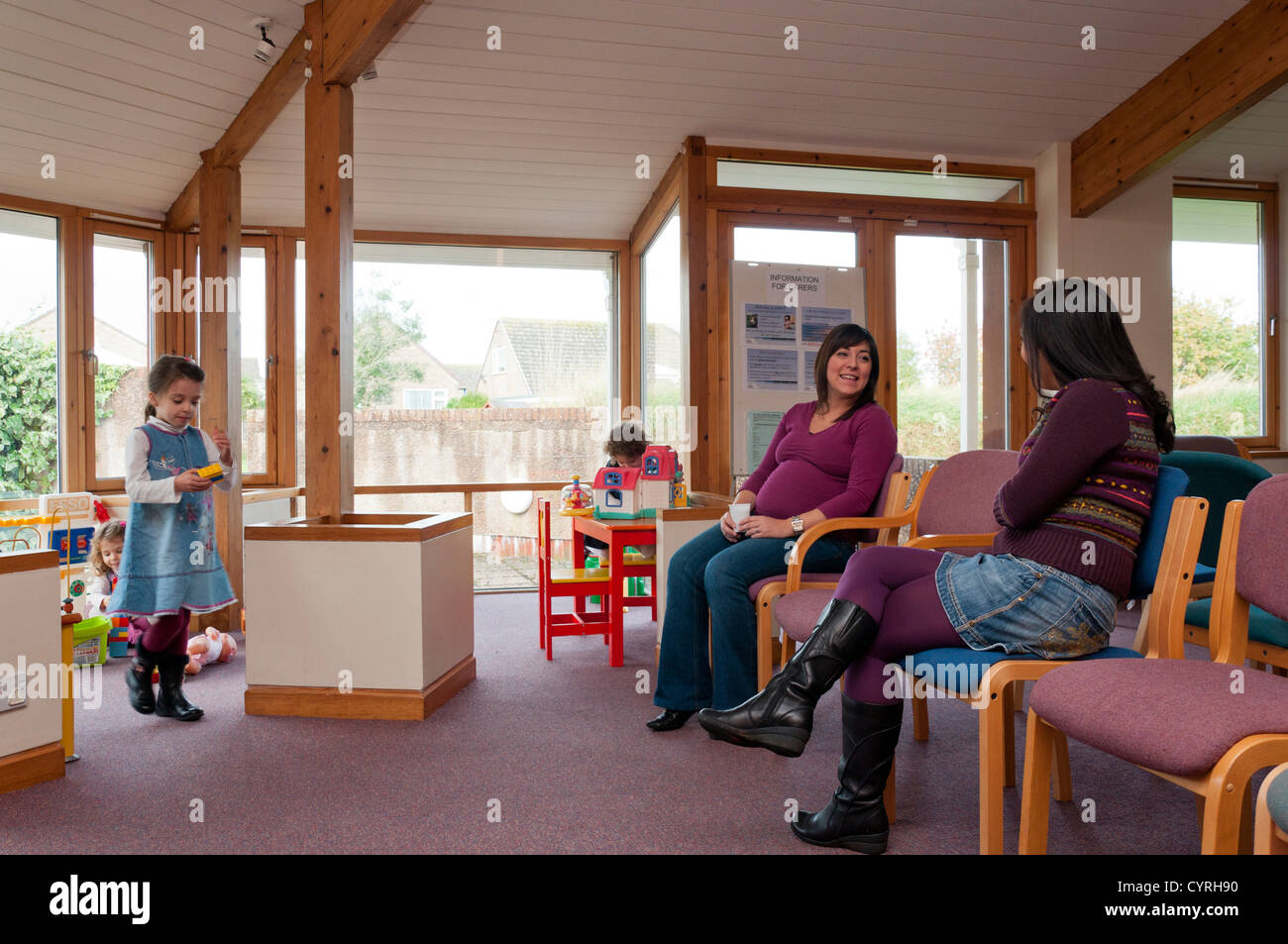 Junge Mütter mit Kindern, die während der Wartezeit in einem Empfangsbereich eine Ärzte-Chirurgie UK chatten. Stockfoto
