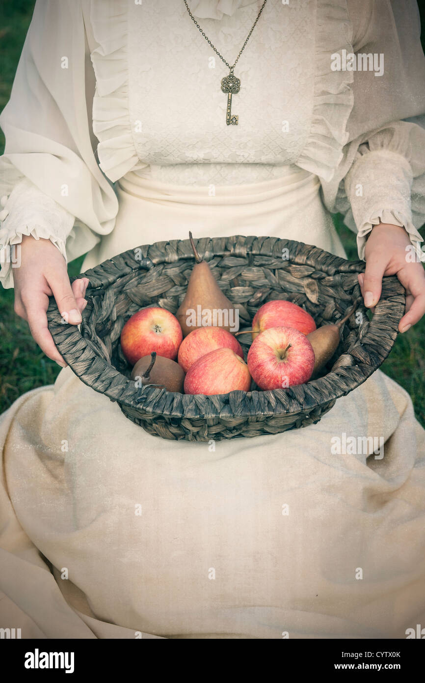 eine Frau in einem weißen edwardian Kleid sitzt auf einer Wiese und einen Korb mit Früchten auf dem Schoß halten Stockfoto
