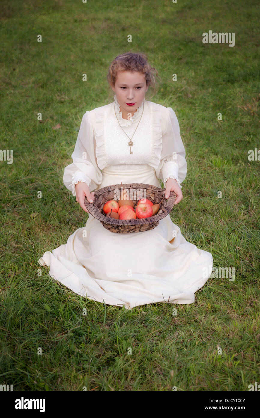 eine Frau in einem weißen edwardian Kleid sitzt auf einer Wiese und einen Korb mit Früchten auf dem Schoß halten Stockfoto