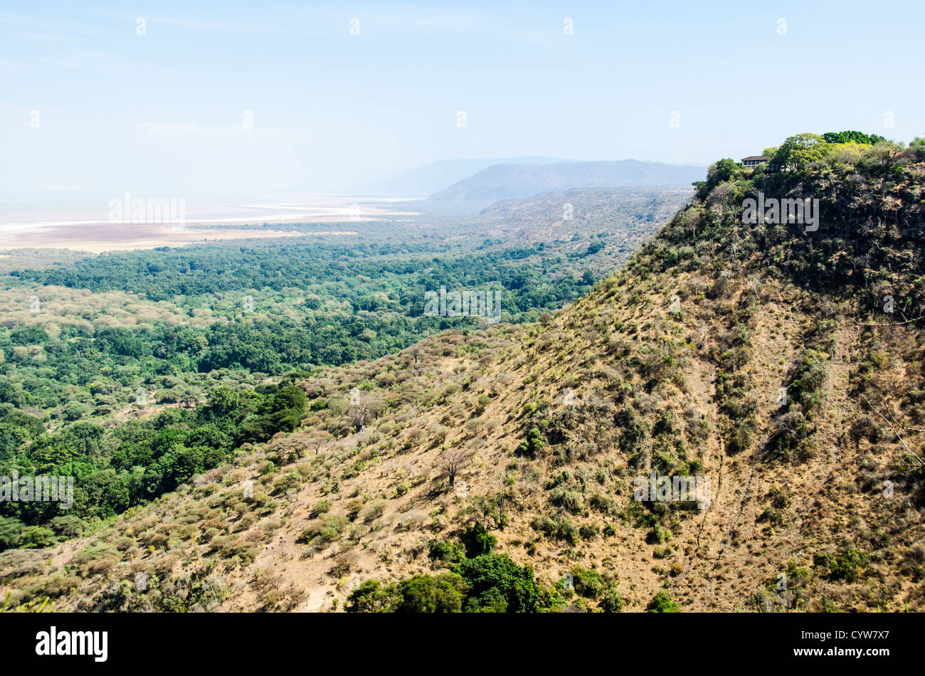 LAKE MANYARA NATIONAL PARK, Tansania – Ein Blick erstreckt sich über den Lake Manyara National Park von einem erhöhten Aussichtspunkt direkt außerhalb der Parkgrenzen. Die erhöhte Perspektive zeigt die weitläufige Landschaft des Parks, mit einem Luxusresort auf der Landzunge auf der rechten Seite. Der Lake Manyara National Park im Norden Tansanias ist Teil des Ostafrikanischen Rift Valley-Systems und dient als wichtiges Naturschutzgebiet. Der Park ist bekannt für seine vielfältigen Ökosysteme, die von alkalischen Seenwässern über Akazienwälder bis hin zu steilen Steilwänden reichen. Der erhöhte Aussichtspunkt bietet Stockfoto