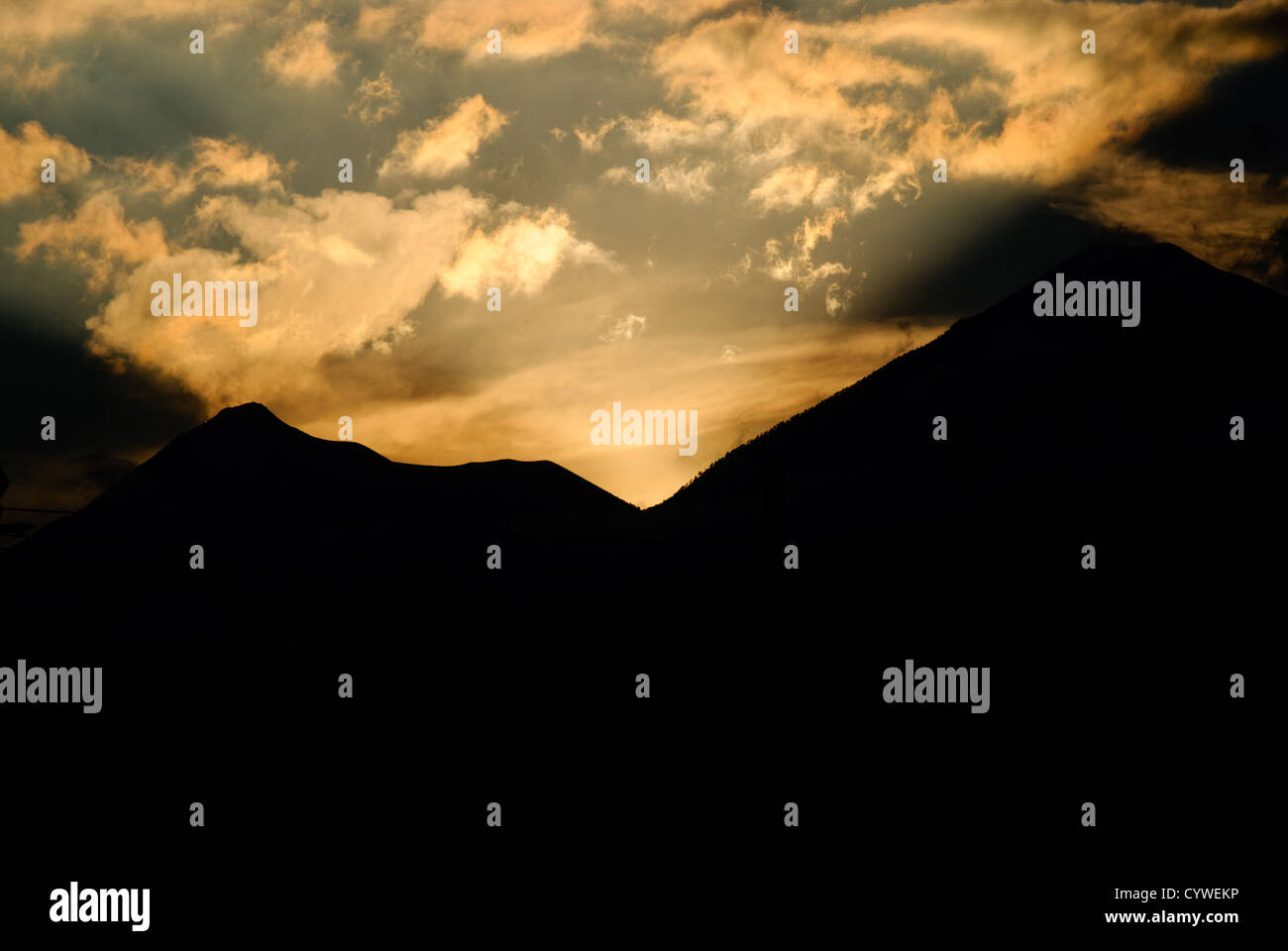 Berge bei Sonnenuntergang Antigua Guatemala // ANTIGUA GUATEMALA, Guatemala — die Sonne untergeht hinter den Bergen um Antigua Guatemala und wirft einen goldenen Glanz auf die Wolken darüber. 1543 als Santiago de Guatemala gegründet, diente die Stadt als Kolonialhauptstadt bis 1773, als sie durch ein Erdbeben weitgehend zerstört wurde. Antigua Guatemala, bekannt für seine gut erhaltene spanische Barockarchitektur und zahlreiche Erdbebenruinen, wurde 1979 zum UNESCO-Weltkulturerbe erklärt. Die Stadt liegt etwa 1.530 Meter über dem Meeresspiegel im zentralen Hochland Guatemalas, umgeben von drei Stockfoto