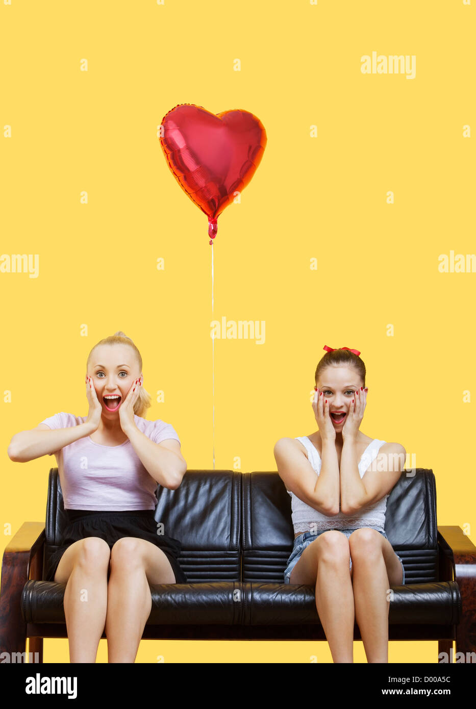 Porträt von zwei begeistert junge Frauen sitzen auf Sofa mit herzförmigen Ballon über gelbem Hintergrund Stockfoto