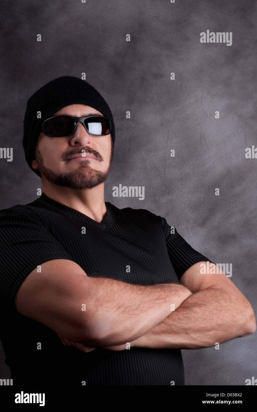 Stolz auf harter Kerl in schwarz. Starke Mann im schwarzen Hemd, Sonnenbrille und Zipfelmütze mit muskulösen Arme verschränkt, Stockfoto