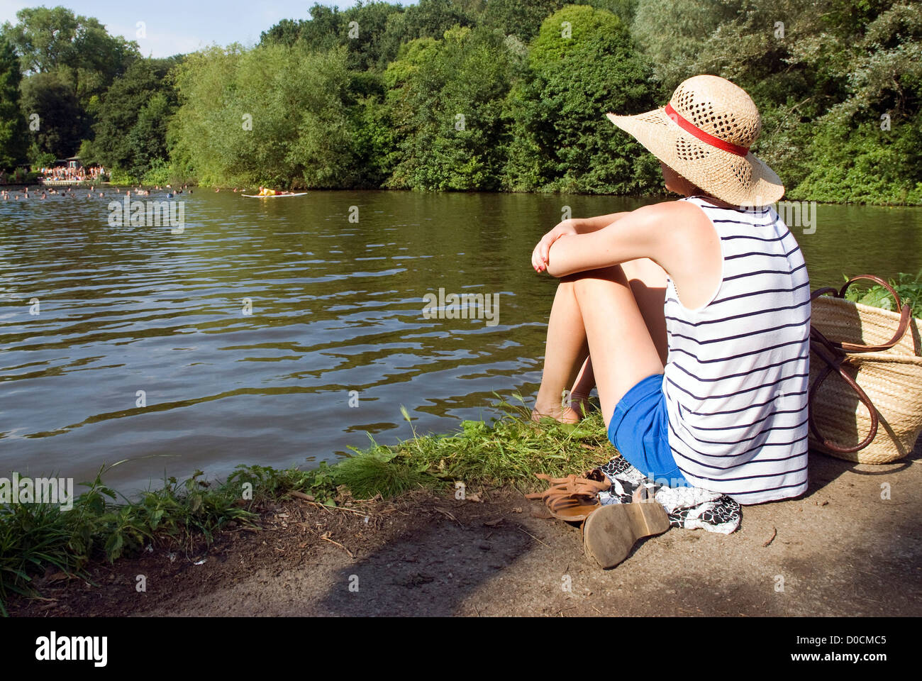 Eine junge Dame sonnt sich in dem breiten Hut an den Ufern des gemischten Teiches auf Hampstead Heath, Nord-London, an einem Sommertag. Stockfoto