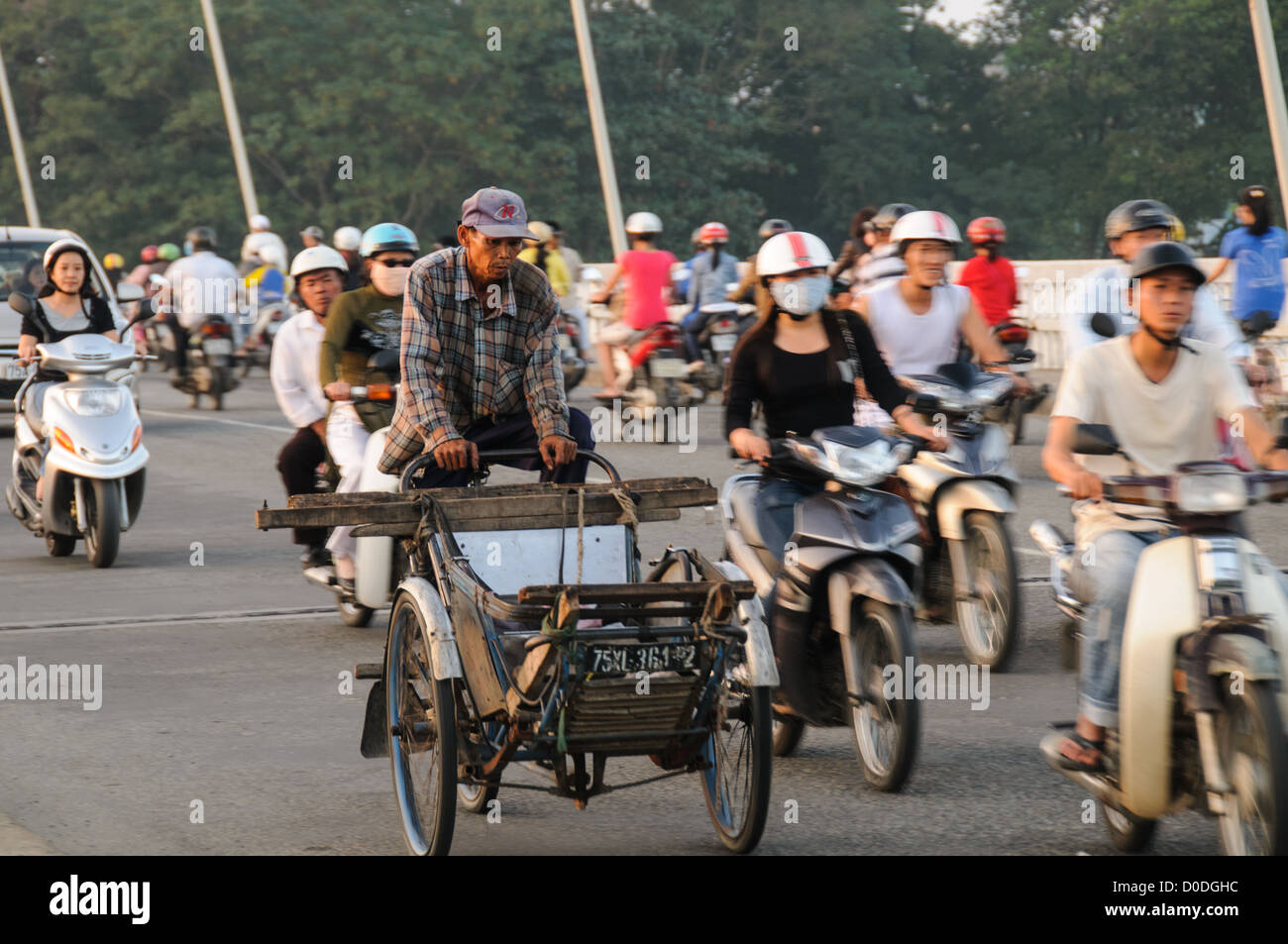 Cargo-Rikscha-Fahrrad-Motorrad-Verkehr Phu Xuan Brücke Hue Vietnam | HUE, Vietnam — Ein Mann tritt ein traditionelles Cargo-Dreirad durch dichten Motorradverkehr auf der Phu Xuan Brücke. Die Brücke, die den Parfümfluss überspannt, dient als wichtige Verbindung zwischen den modernen südlichen Bezirken von Hue und dem nördlichen Ufer, wo die im 19. Jahrhundert erbaute Kaiserstadt — Sitz der Nguyen-Dynastie von 1802 bis zur Abdankung Bao Dais 1945 — den UNESCO-Weltkulturerbestätten-Komplex von Hue verankert. Das pedalbetriebene Pritschenrad, historisch der Rückgrat der vietnamesischen Stadtlogistik, konkurriert nun Stockfoto