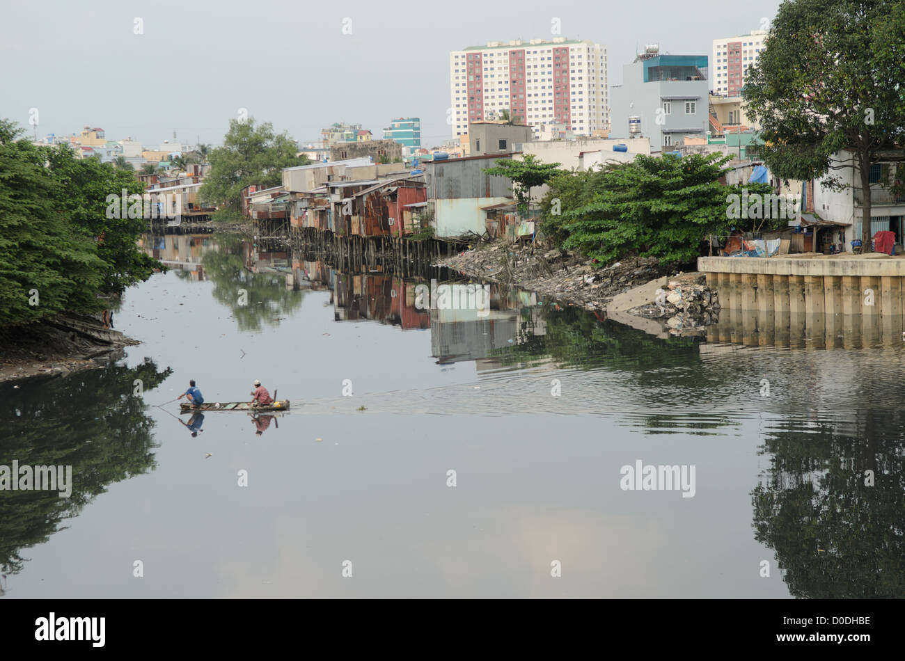 Angeln vom Kanu Saigon-Fluss Ho-Chi-Minh-Stadt Vietnam // HANOI, Vietnam — zwei Männer in einem kleinen Holzkanu fischen mit einem Netz in den stillen Gewässern eines Nebenflusses des Saigon-Flusses in Ho-Chi-Minh-Stadt, Hanoi, mit provisorischen Häusern am Ufer und hohen Hochhäusern im Hintergrund. Stockfoto