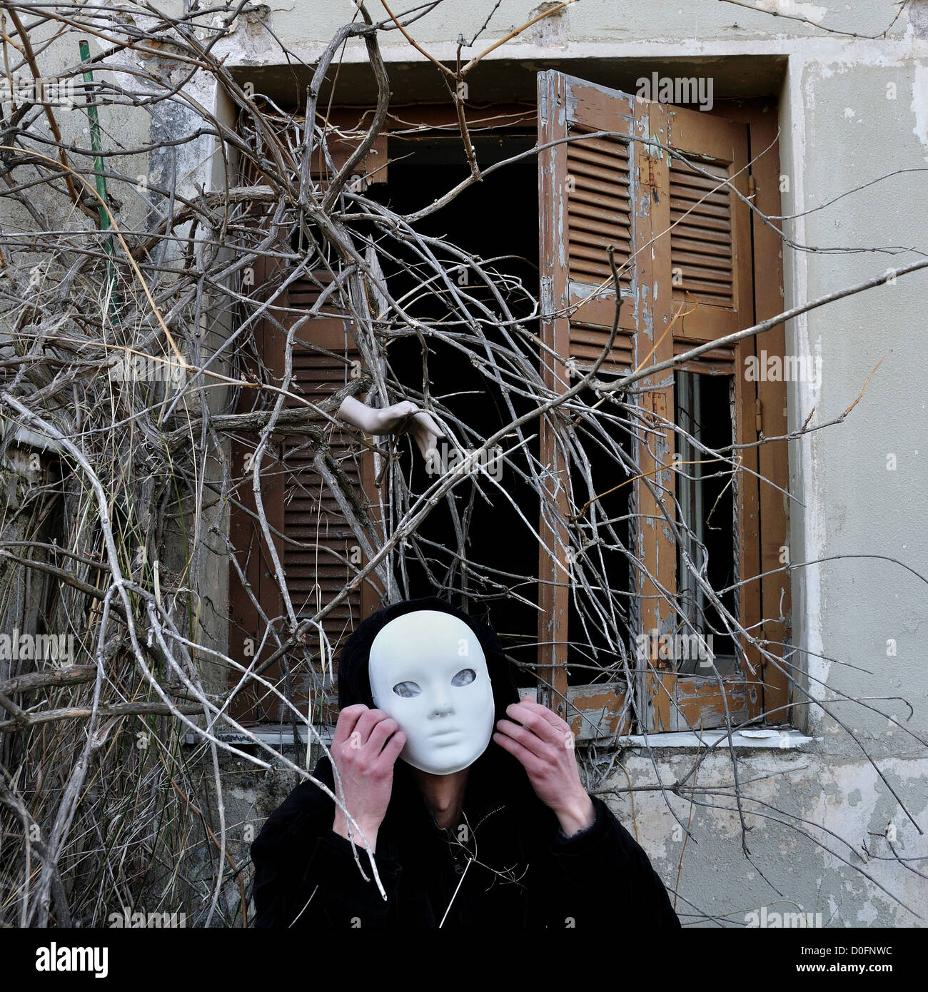 Groteske weiße Maske Figur und Spukhaus Fenster mit verworrenen verwilderten Pflanzen und gruselige Dummy-Puppe Hand. Stockfoto