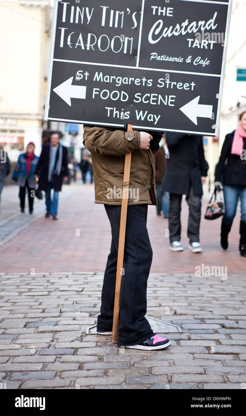 Ein Mann stand in der High Street hält ein Plakat mit Wegbeschreibungen für lokale Geschäfte. Stockfoto