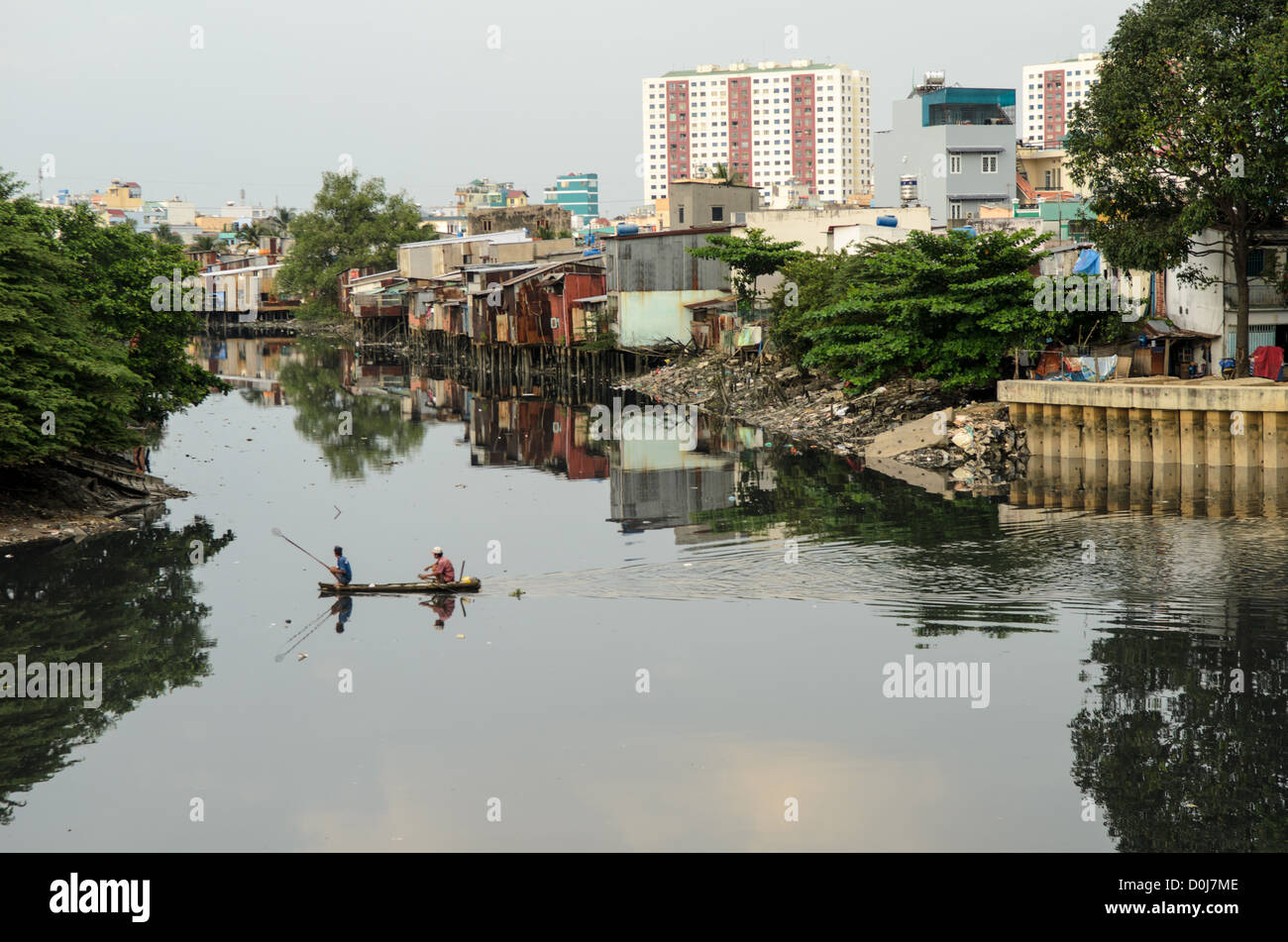 Angeln vom Kanu Saigon Fluss Ho Chi Minh Stadt // HANOI, Vietnam — zwei Männer in einem kleinen Holzkanu fischen mit einem Netz in den stillen Gewässern eines Nebenflusses des Saigon Flusses in Ho Chi Minh Stadt, Hanoi, mit provisorischen Häusern am Ufer und hohen Hochhäusern im Hintergrund. Stockfoto