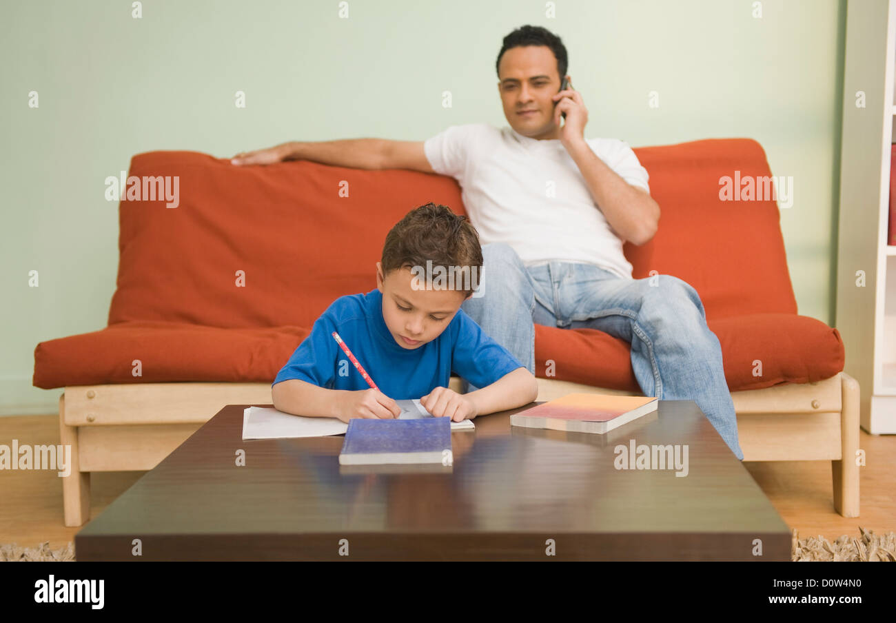 Boy Hausaufgaben mit Vater sprechen auf einem Handy hinter ihm Stockfoto