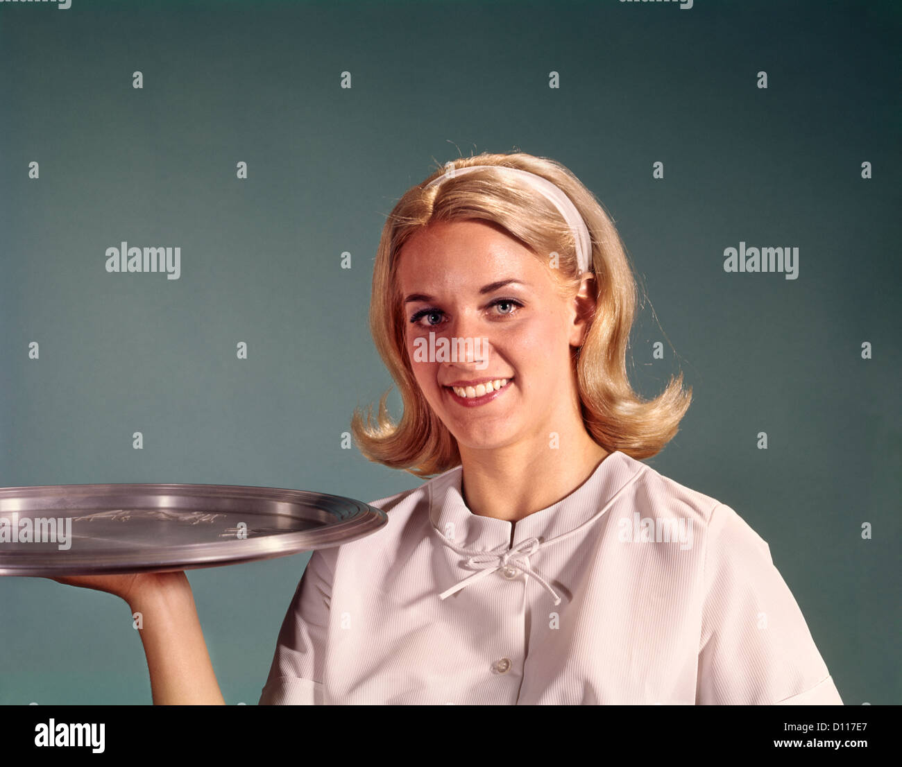 1960ER JAHRE BLONDE FRAU KELLNERIN HOLDING SERVIERTABLETT BLICK AUF DIE KAMERA ZU LÄCHELN Stockfoto