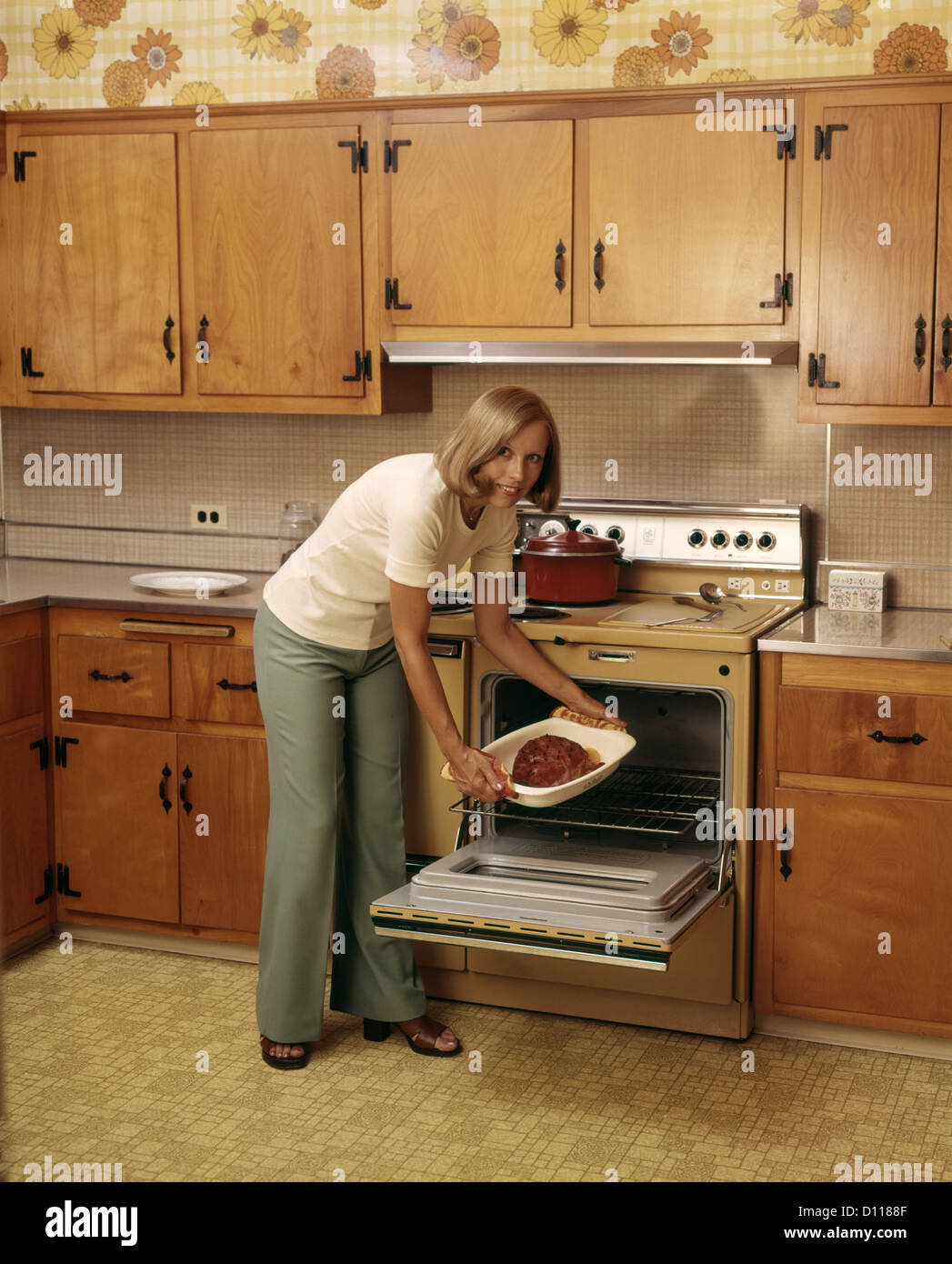 1970ER JAHREN FRAU NEHMEN SCHINKEN AUS KÜCHE OFEN GEBACKEN, BLICK IN DIE KAMERA Stockfoto