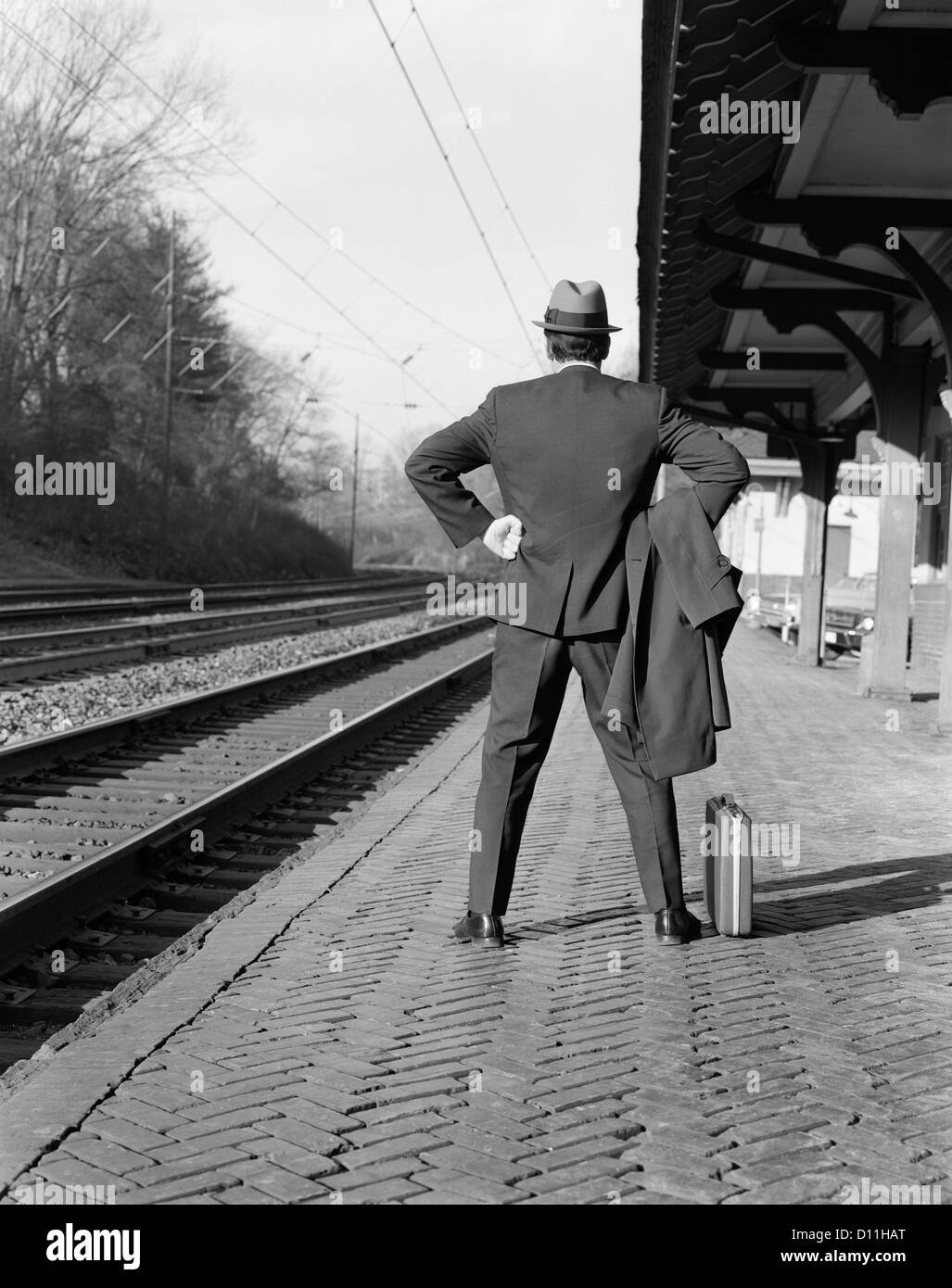 1950S 1960S MANN PENDLER ZURÜCK ANZEIGEN STEHEND HÄNDE AUF DEN HÜFTEN AKTENTASCHE ZUG WARTEN ODER EINEN ZUG VERPASST Stockfoto