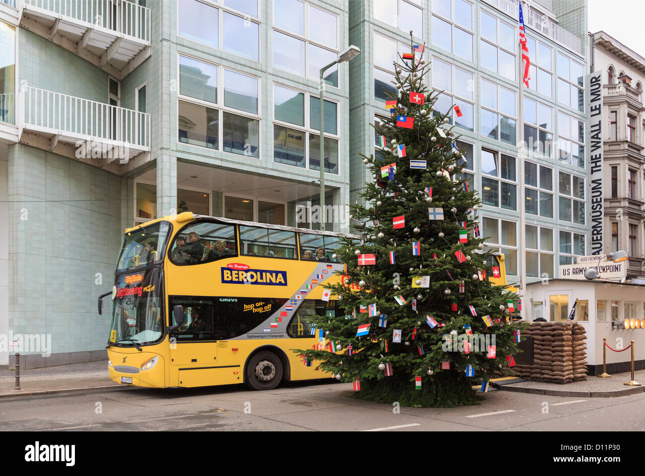 Touristischen Sightseeingbus vorbei Checkpoint Charlie mit einem Weihnachtsbaum dekoriert mit internationalen Fahnen in Berlin Deutschland Stockfoto