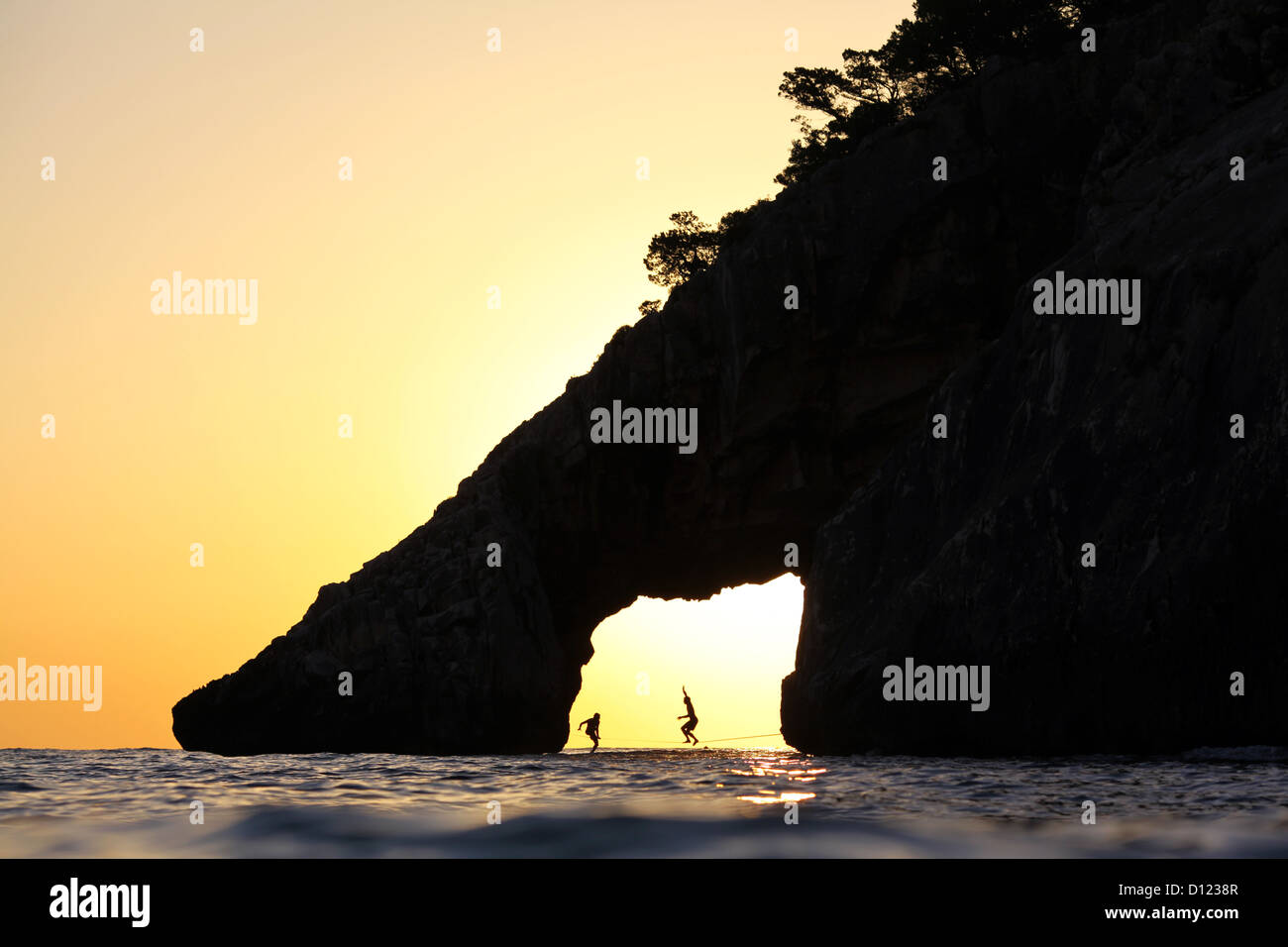 Zwei Slackliner zu Fuß auf einer Slackline in einer offenen Höhle am Wasser in Cala Goloritze, Sardinien, Italien, bei Sonnenuntergang. Stockfoto