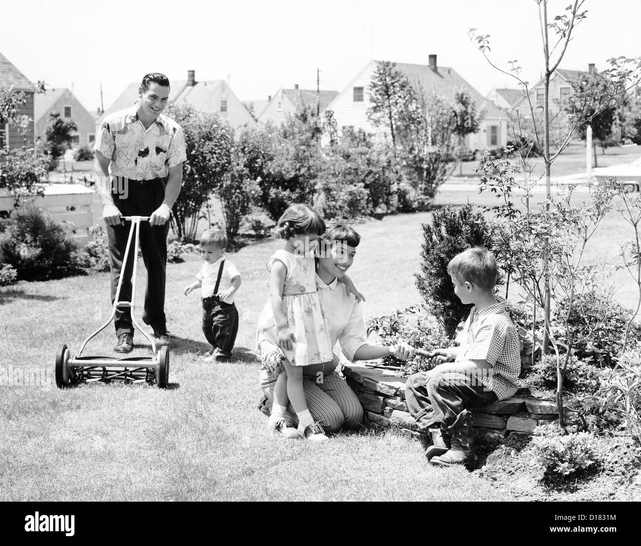 Mann mäht den Rasen, während Frau mit Kindern spielt Stockfoto