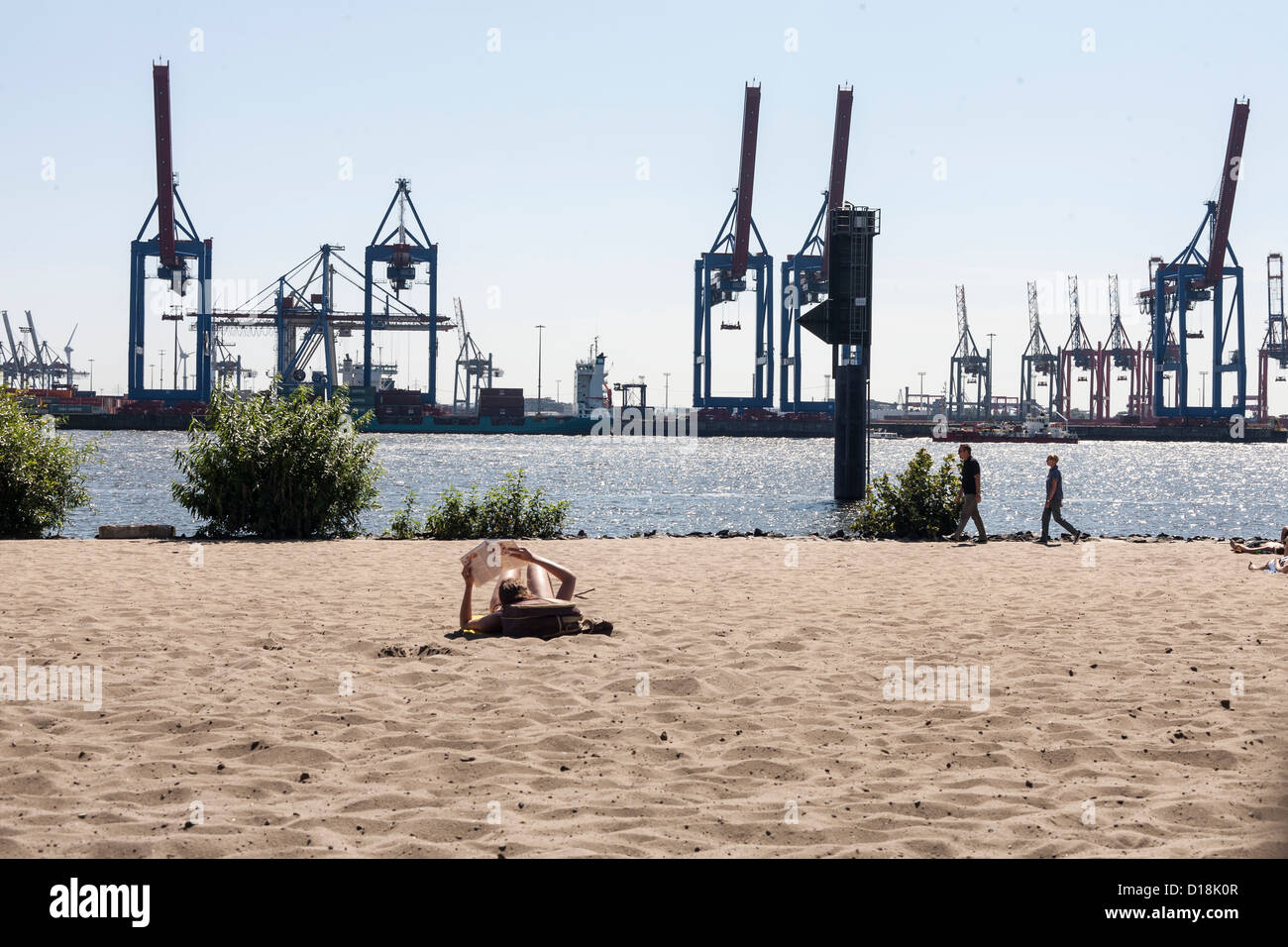 Frau liest Zeitung auf Mann machte Strand in Hamburg mit Industriehafen Hintergrund Stockfoto