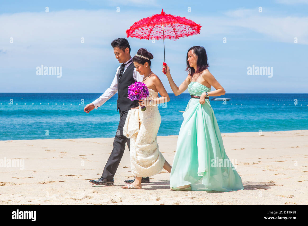 Hochzeitsfeier am Pansea Beach, Surin Beach, Phuket, Thailand - Braut, Bräutigam und Brautjungfer mit roten Sonnenschirm Stockfoto