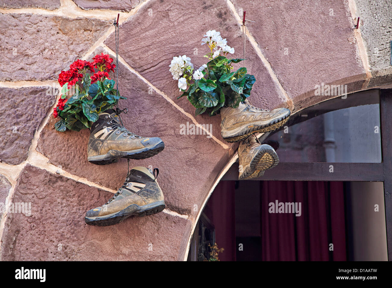 Alte Wanderschuhe, die über dem Eingang mit Blumen in ihnen hängen, entlang des Camino de Santiago de Compostela, Stockfoto