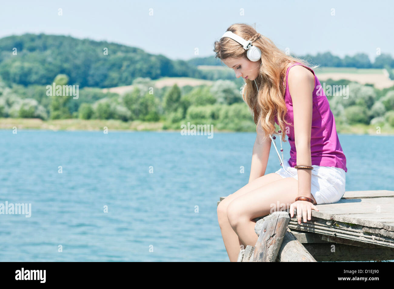 Blonde junge Frau sitzt mit Kopfhörern auf einem Steg am See Stockfoto