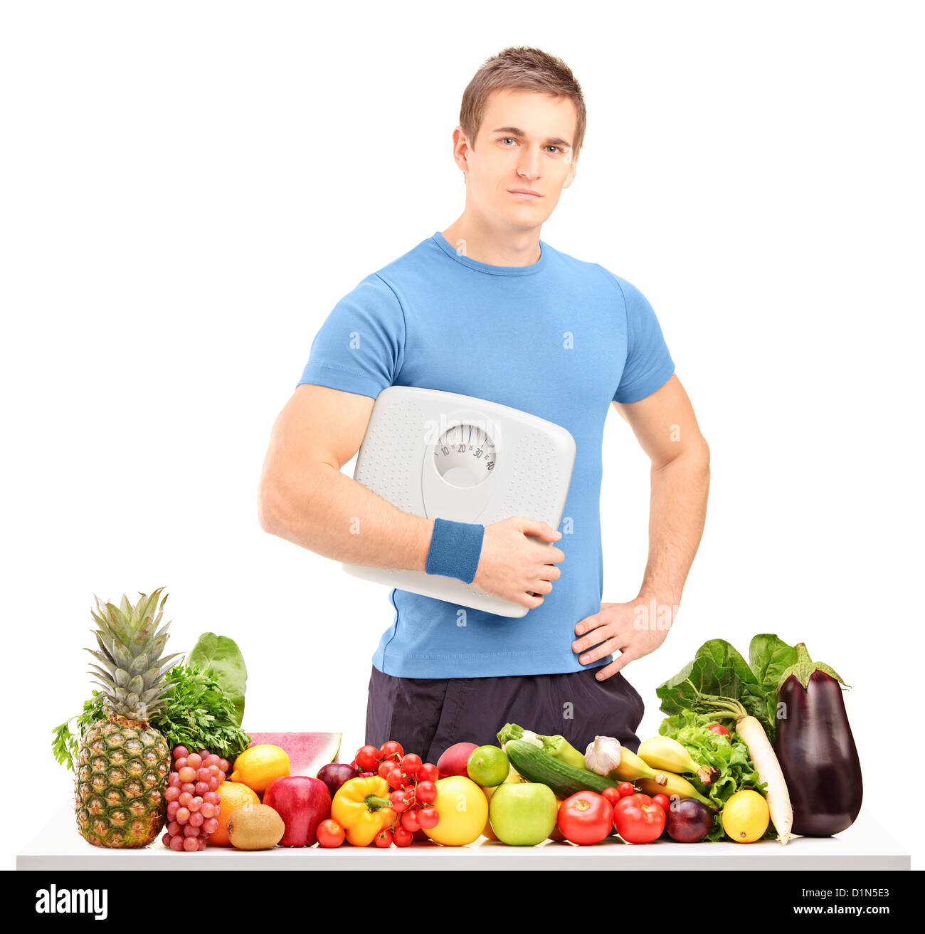Ein männlicher Athlet hält eine Waage hinter einem Tisch voller Obst und Gemüse isoliert auf weißem Hintergrund Stockfoto