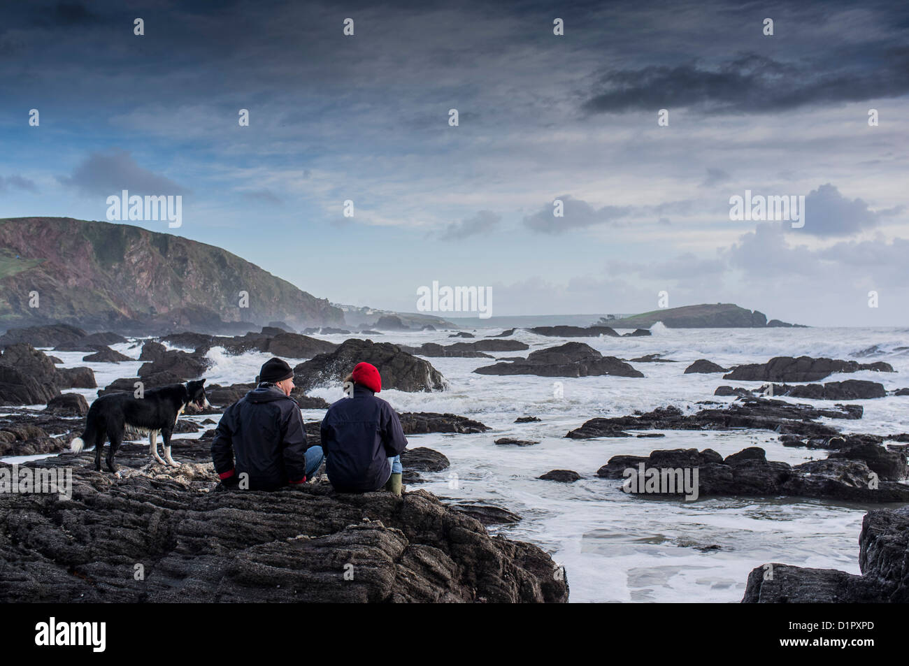 Pärchen und Hund beobachten den Sonnenuntergang über dem Meer von einem felsigen Strand in Richtung Burgh Island. Ayrmer Cove, Devon. GROSSBRITANNIEN Stockfoto
